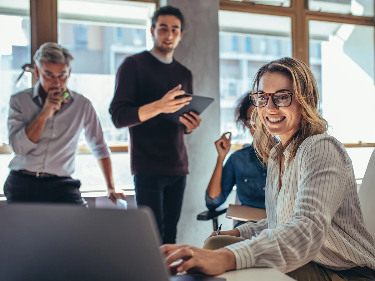 AdobeStock_265815625-crop A smiling woman wearing glasses is focused on her work while using a laptop.