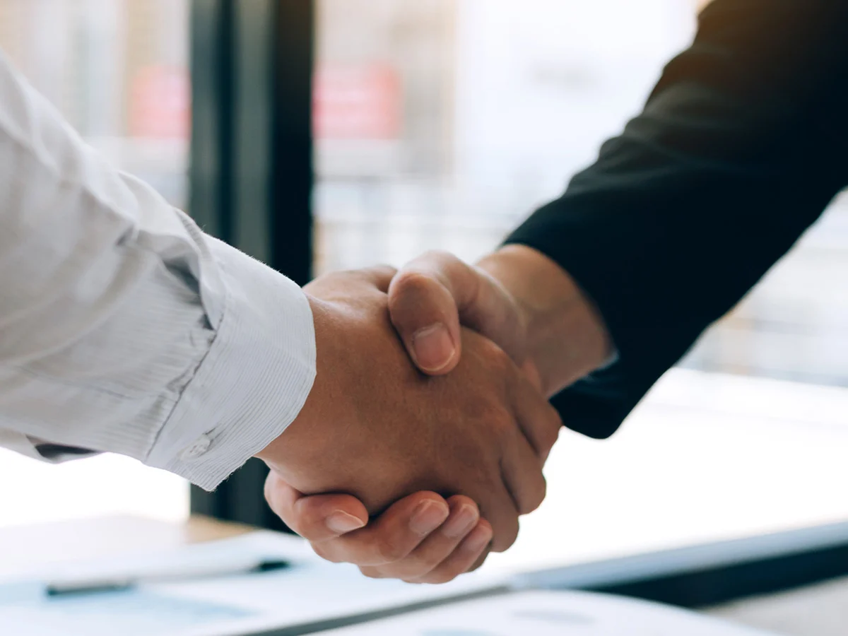 AdobeStock_283138003-crop Two business professionals shaking hands in front of a large window, symbolizing a successful partnership or agreement.